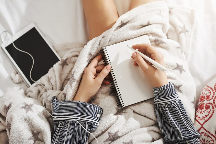 Foto grátis não há lugar como a cama. mulher criativa e descontraída, deitado sob o cobertor, ouvindo música em fones de ouvido via tablet digital, fazendo anotações enquanto trabalha em casa. menina usa imaginação