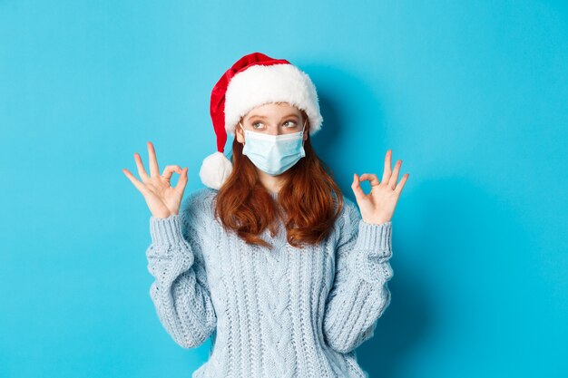 Natal, quarentena e conceito covid-19. Linda garota ruiva adolescente com chapéu de Papai Noel e suéter, usando máscara de coronavírus, mostrando sinais de ok, aprovar e elogiar algo.