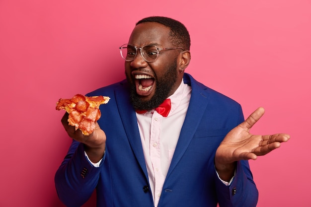 Negro faminto morde um pedaço enorme de pizza, tem apetite, usa roupas formais e posa de óculos contra o espaço rosa