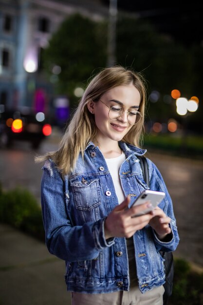 Noite ao ar livre da jovem mulher usando a cara do telefone inteligente iluminada pelo screenlight. Internet, rede social, conceito de tecnologia