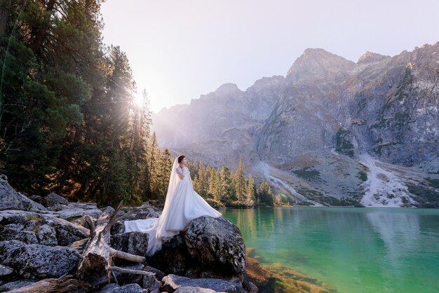 Noiva atraente está de pé sobre a rocha com uma vista deslumbrante do lago das montanhas com água de cor verde no dia ensolarado, montanhas Tatry