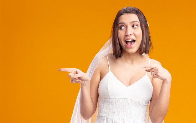 Noiva em lindo vestido de noiva, parecendo feliz e alegre, sorrindo amplamente apontando com o dedo indicador para o lado em pé sobre a parede laranja