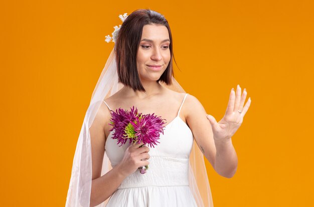 Noiva em um lindo vestido de noiva com buquê de flores olhando para o anel em seu dedo em pé sobre uma parede laranja