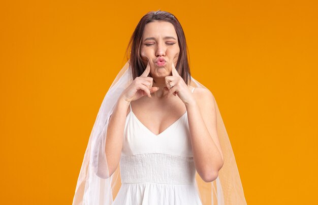 Noiva em um lindo vestido de noiva soprando as bochechas apontando com o dedo indicador para eles em pé sobre a parede laranja