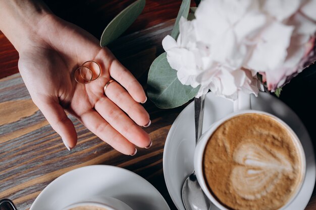 Noiva segura dois anéis de casamento de ouro no braço dela antes de duas xícaras com café