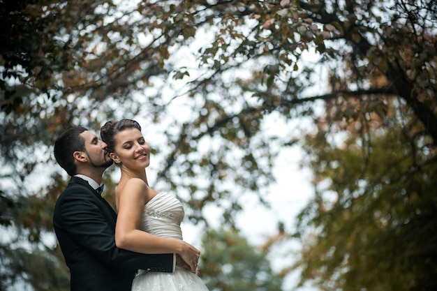 Foto grátis noivo abraça gentilmente a noiva com um buquê por trás em um parque