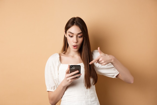 Notícia empolgante no telefone surpreendeu a mulher bonita lendo promoção online na tela apontando para smartphone e ...