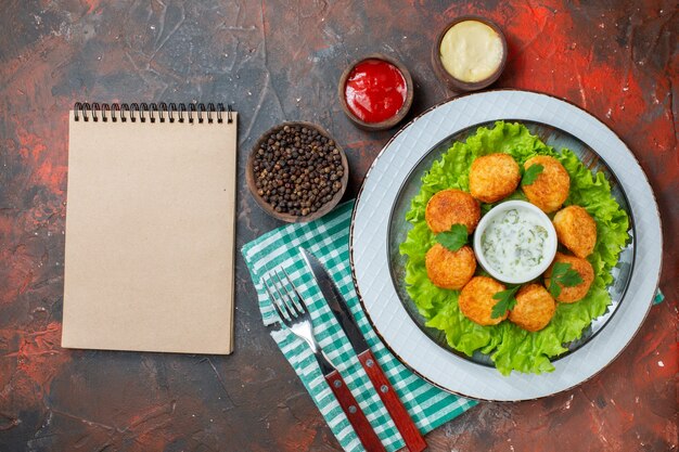 Nuggets de frango de cima, alface e molho no prato, molhos e pimenta preta em tigelas, caderno de garfo e faca na mesa escura