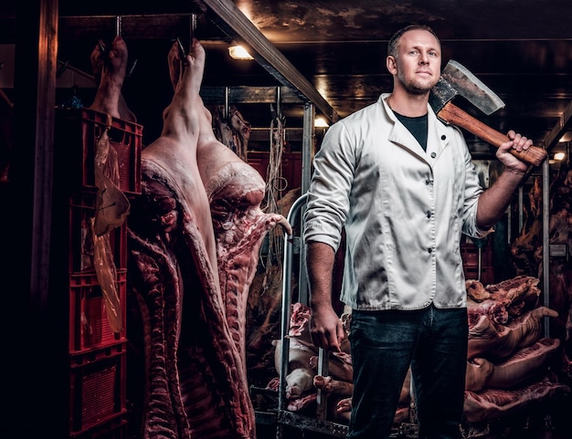 O açougueiro em uma camisa branca de trabalho segurando o machado em um armazém refrigerado no meio de carcaças de carne