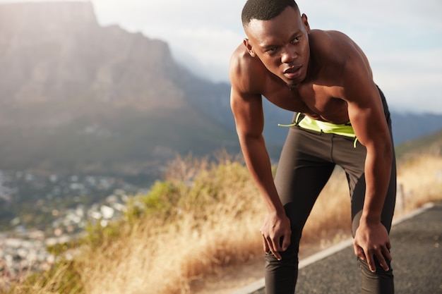 O adulto negro e saudável faz exercícios em estradas de montanha, treina para a maratona, mantém as duas mãos nos joelhos, olha pensativo para a distância, corre no campo, tem expressão facial determinada.