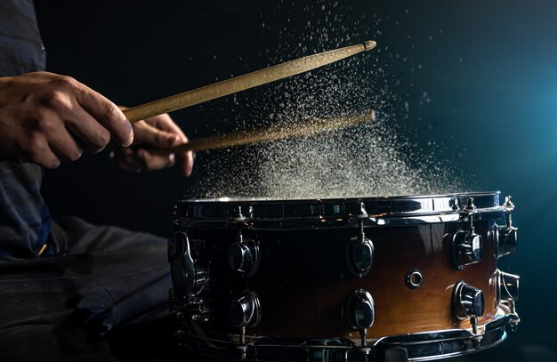 O baterista usando baquetas batendo na tarola com salpicos de água no fundo preto sob a iluminação do estúdio close-up.