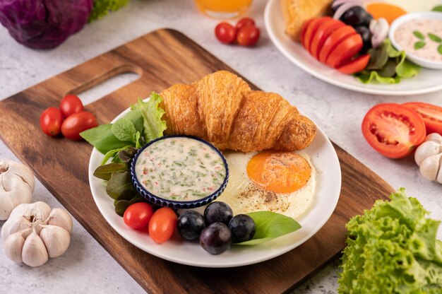 O café da manhã consiste em croissant, ovo frito, molho para salada, uvas pretas e tomate.