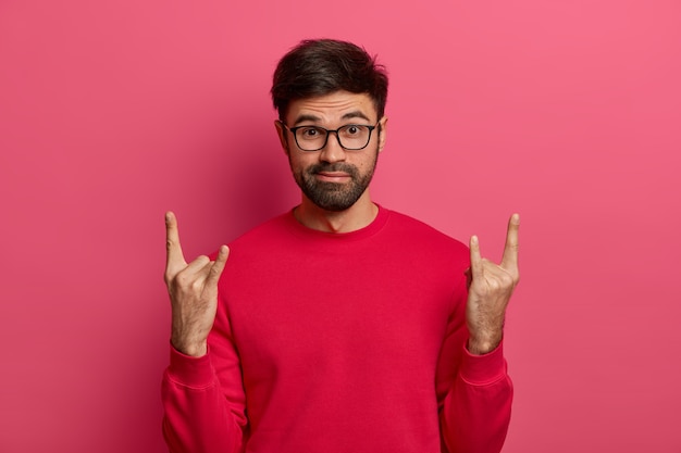 Foto grátis o cara barbudo espantado visita um festival de música incrível, faz gestos de rock n roll, se diverte ouvindo a música favorita de meatal pesado, vestido casualmente, posa contra a parede rosa. rock vive para sempre