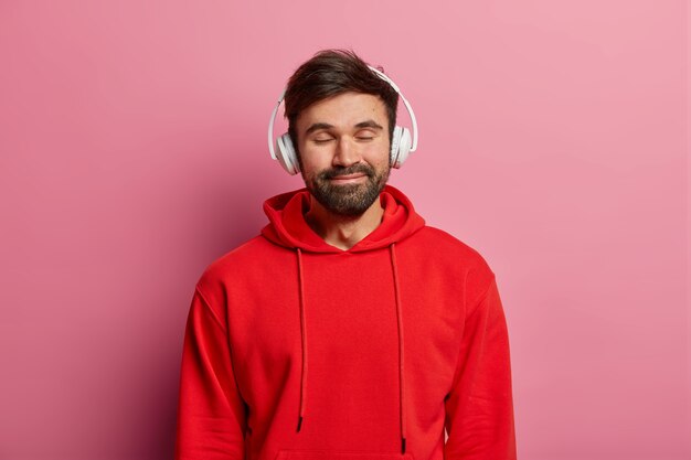 O cara barbudo satisfeito gosta de ouvir música em fones de ouvido estéreo, fecha os olhos e sorri gentilmente, usa um moletom vermelho, se sente bem, é modelo em uma parede rosa pastel. Adolescentes, hobby, conceito de estilo de vida