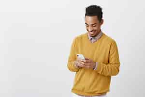 Foto grátis o cara conheceu uma garota engraçada na rede social. retrato de um homem afro-americano feliz e satisfeito, sorrindo amplamente enquanto olha para a tela do smartphone, digitando uma mensagem ou assistindo um vídeo hilário