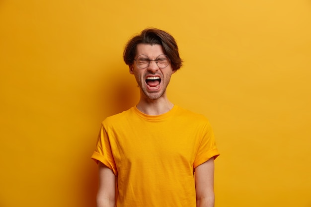 O cara emocional fica com a boca bem aberta, grita de decepção, grita com os olhos fechados, sente-se perturbado porque perdeu uma aposta enorme, vestido com uma camiseta amarela brilhante, perde a paciência, tem problemas de vida