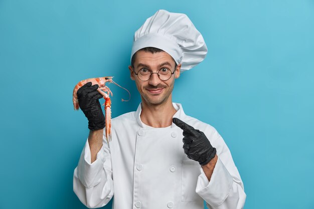 O chef cozinheiro positivo aponta para lagosta cozida, usa luvas pretas de borracha, uniforme branco