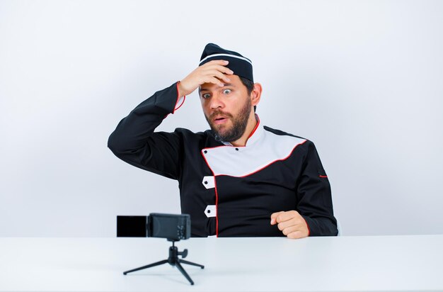 O chef do blogueiro surpreso está colocando a mão na frente sentando-se na frente de sua mini câmera no fundo branco