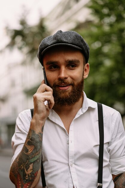 O close-up do retrato do jovem homem tatuado barbudo chama ao telefone na rua. Um cara romântico em uma camisa branca, boné e suspensórios. Peaky Blinders. antiquado, retrô.