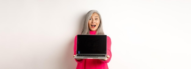 Foto grátis o conceito de comércio eletrônico surpreendeu a mulher sênior asiática mostrando a tela do laptop em branco e parecendo uma demonstração feliz