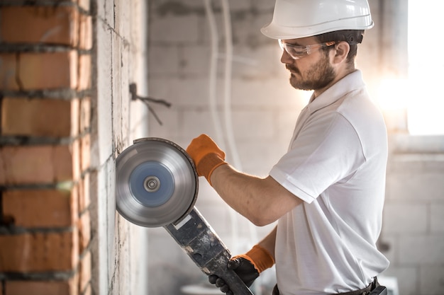 O construtor industrial trabalha com uma rebarbadora profissional para cortar tijolos e construir paredes interiores. Eletricista.