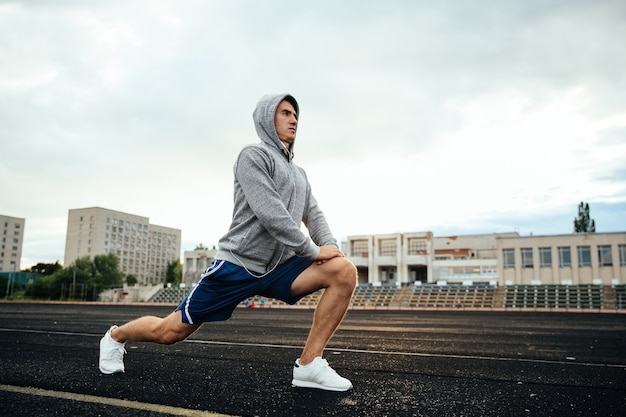 O desportista novo considerável que faz lunges, exercícios, escutando a música, no estádio.