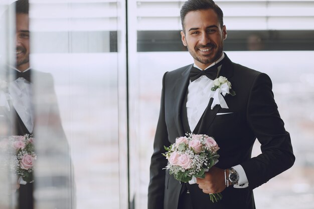 O elegante e elegante noivo está no quarto do hotel com um buquê de flores