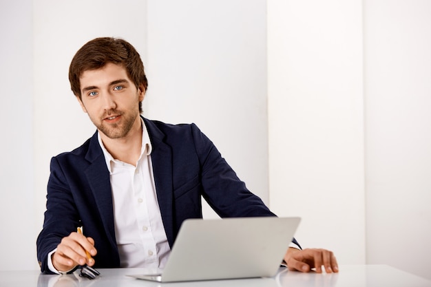 O empresário masculino considerável no terno senta-se na mesa de escritório com laptop, olha satisfeito