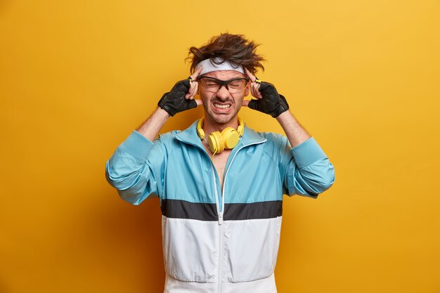 O esportista frustrado sente uma dor de cabeça insuportável após o treino, mantém as mãos nas têmporas, cerra os dentes, concentra-se na tarefa, usa luvas esportivas e roupa ativa, sofre de enxaqueca. Cara esportivo doente e cansado