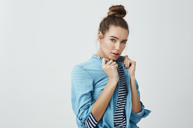 O estilo está no sangue dela. Foto de estúdio do modelo feminino europeu bonito, puxando a camisa jeans enquanto usava mais de camiseta listrada, olhando sensualmente e com confiança