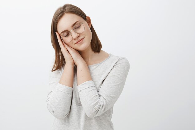 O estudante Harworking desejava travesseiro e sono. Charmosa garota sonhadora e inteligente de óculos e blusa branca inclinando a cabeça nas palmas das mãos sorrindo gentilmente e ternamente com os olhos fechados como se estivesse dormindo e tendo sonhos