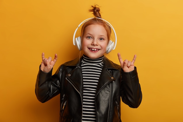 Foto grátis o garoto roqueiro otimista faz sinais de trompa com os dedos, gosta de ouvir heavy metal em fones de ouvido, tem um nó de cabelo sexy, usa jaqueta de couro, sente-se animado e radiante, relaxa dentro de casa