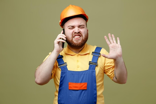 O gesto tenso mostrando olá fala ao telefone jovem construtor de uniforme isolado em fundo verde
