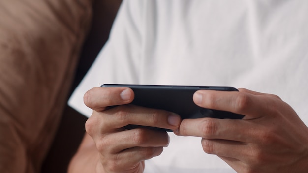 O homem asiático novo que usa o telefone celular que joga videogames na televisão na sala de visitas, sentimento masculino feliz usando relaxa o tempo que encontra-se no sofá em casa. Os homens jogam jogos relaxam em casa.