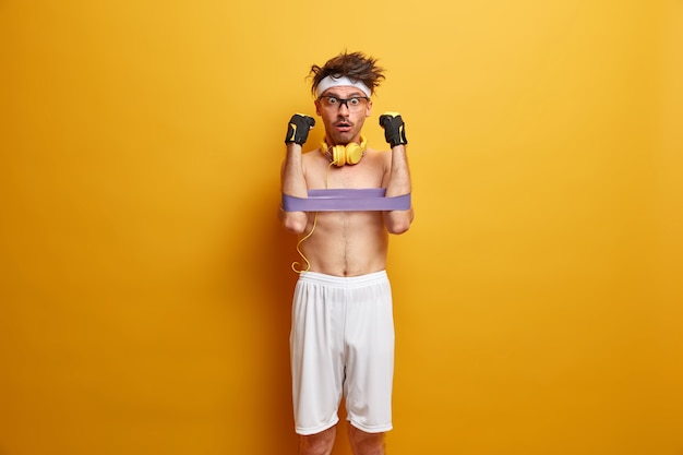 Foto grátis o homem atleta desenvolve os músculos das mãos com goma de mascar, encara com expressão de choque, usa bermuda branca, fita para a cabeça, usa fones de ouvido estéreo, isolados na parede amarela. fique forte e saudável