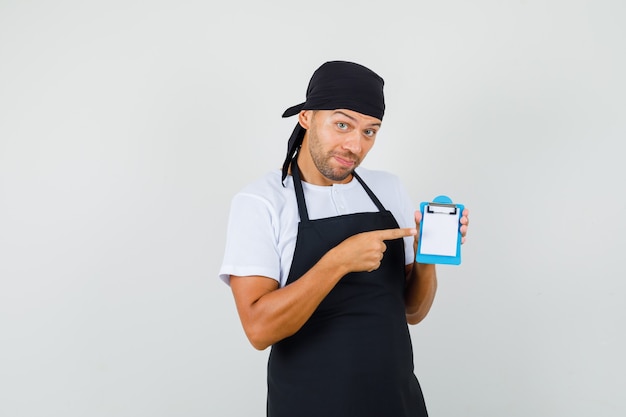 O homem Baker apontando para a mini prancheta em camiseta, avental e parecendo otimista