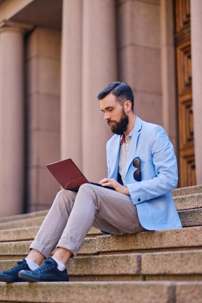 O homem barbudo elegante senta-se em um degrau e usando o laptop.