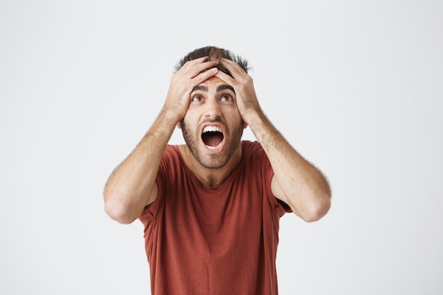 Foto grátis o homem barbudo latino-americano de camisa vermelha reage expressivamente às más notícias do trabalho ficando irritado com seu chefe. infeliz cara gritando sendo demitido do trabalho.