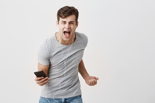 O homem bonito e emocionado parece excitado, grita ou grita, ouve a faixa de áudio com fones de ouvido e franze a testa. Cara jovem muscular usa tecnologia moderna