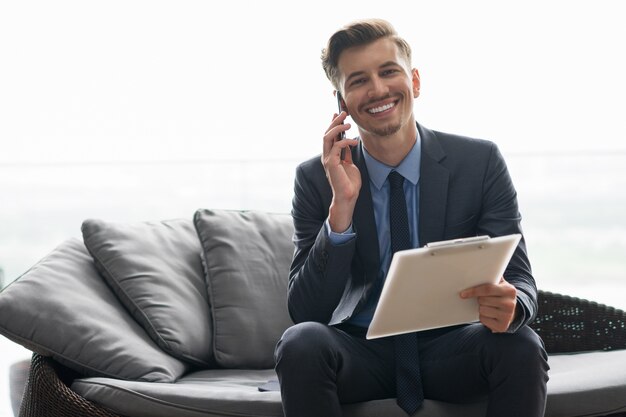 O homem de negócios que convida o telefone no sorriso do sofá