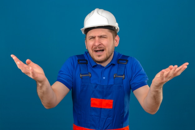 O homem do construtor que veste o uniforme da construção e o capacete de segurança que discutem têm a discussão que levanta as mãos em consternação que encolhe os ombros e confundido com a face incomodada irritada sobre o azul isolado