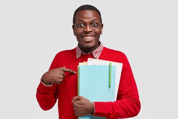 O homem feliz de pele escura aponta para si mesmo, tem expressão feliz, sorriso largo, carrega livro didático, pergunta se ele realmente merece nota excelente