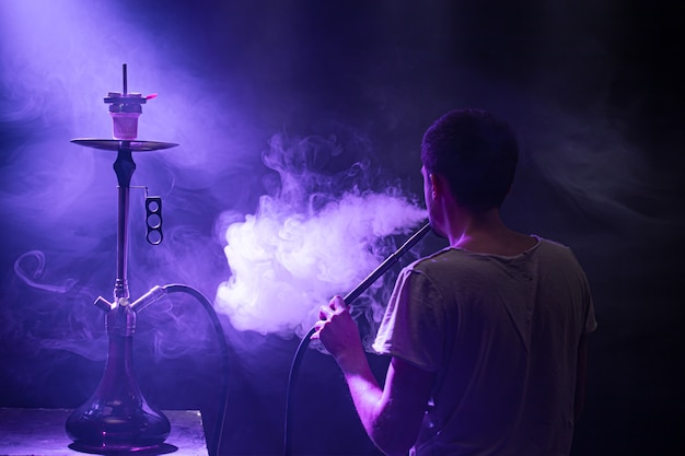 Foto grátis o homem fumando o clássico shisha. lindos raios coloridos de luz e fumaça. o conceito de fumar narguilé.
