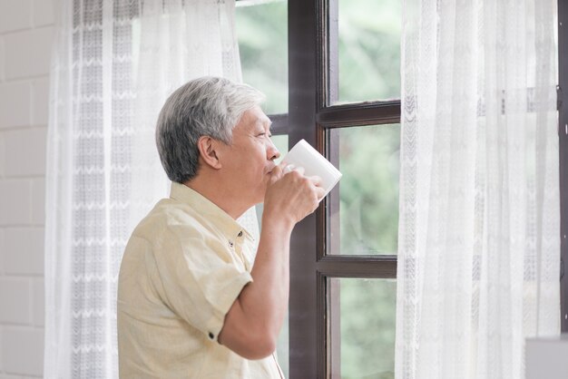O homem idoso asiático feliz que sorri e que bebe uma xícara de café ou um chá perto da janela na sala de visitas, homem superior de Ásia abre as cortinas e relaxa na manhã.