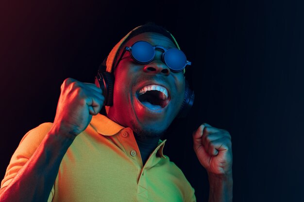 O homem jovem bonito feliz hipster ouvindo música com fones de ouvido no estúdio preto com luzes de néon. Discoteca, boate, estilo hip hop, emoções positivas, expressão facial, conceito de dança