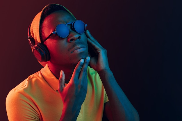 O homem jovem bonito sério triste hipster ouvindo música com fones de ouvido no estúdio preto com luzes de néon.