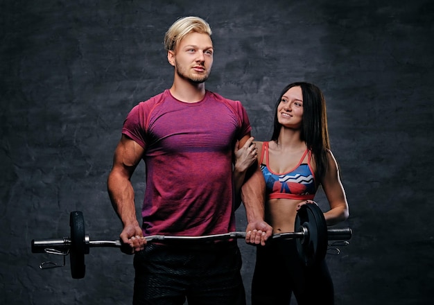 O homem loiro e atlético detém peso de barra e modelo feminino de fitness magro sobre fundo cinza escuro.