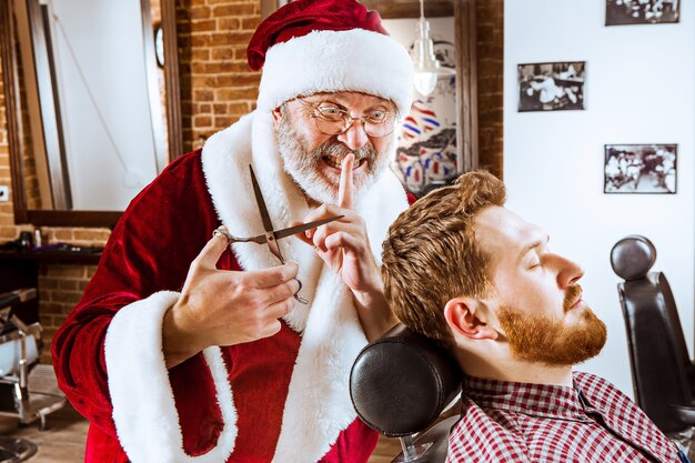O homem sênior com fantasia de Papai Noel trabalhando como mestre pessoal com uma tesoura na barbearia antes do Natal