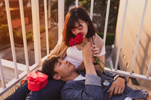 O homem sorrindo deitado sobre as pernas de uma mulher com um buquê de rosas