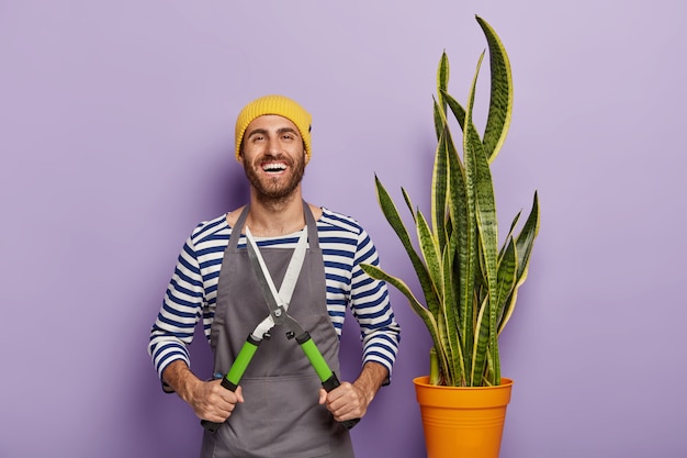 O jardineiro sorridente segura uma tesoura de poda, se preocupa com a planta de cobra no vaso, usa chapéu e avental, tem uma expressão alegre, sendo florista profissional